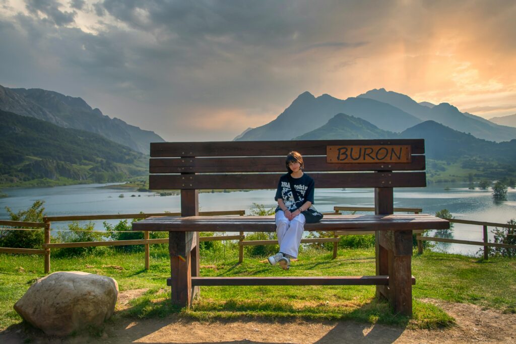 A young girl sits peacefully on a bench by the lake in Burón, surrounded by mountains