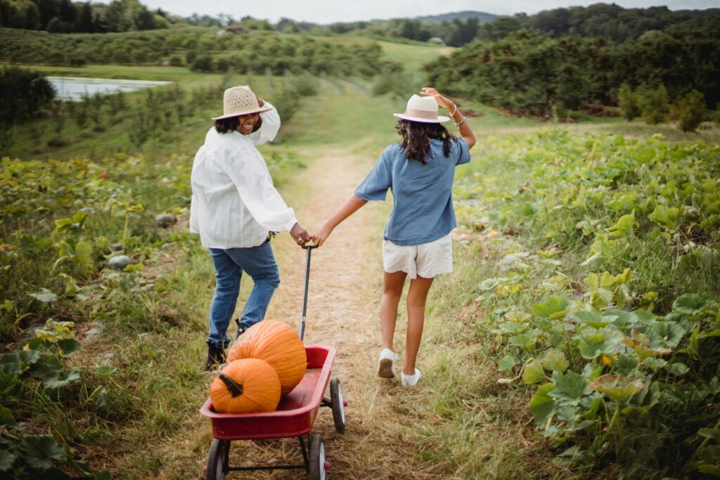 Two young women haul a wheelbarrow full of pumpkins down a lush green path: supporting a new generation of agricultural entrepreneurship and sustainable local produce.