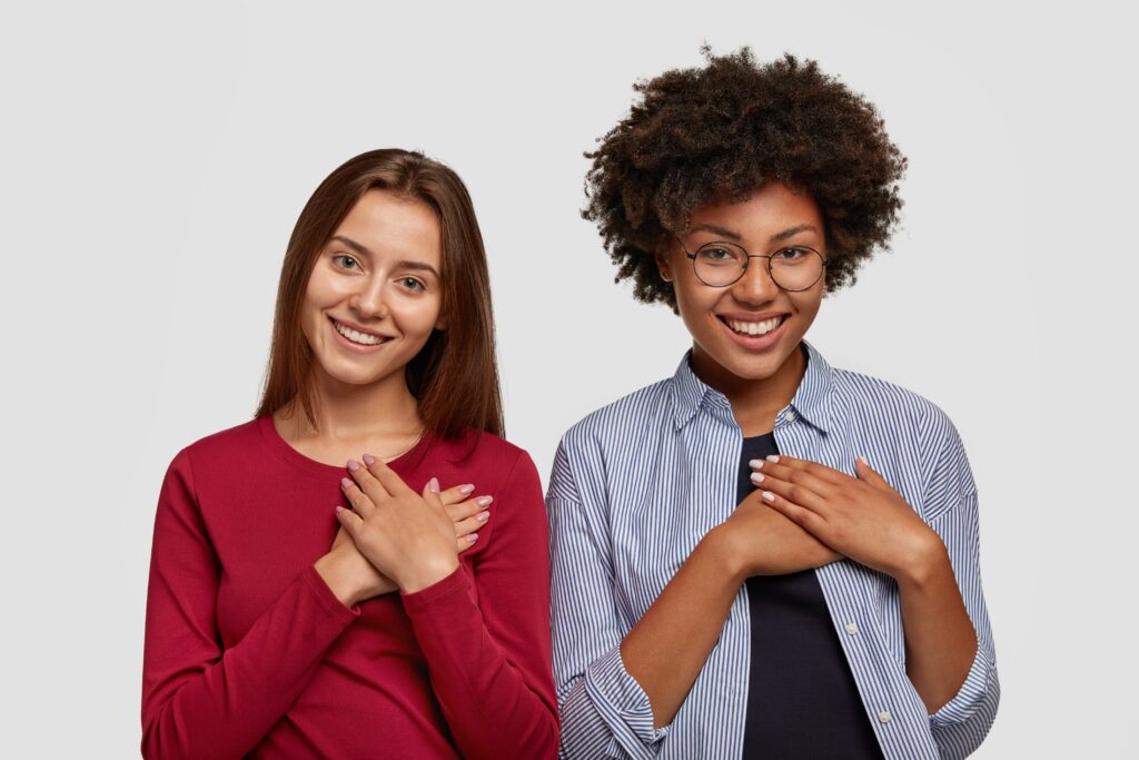 Two young women in Barcelona share a warm, genuine smile, each with a hand placed over their heart in a gesture of sincerity and friendly connection.