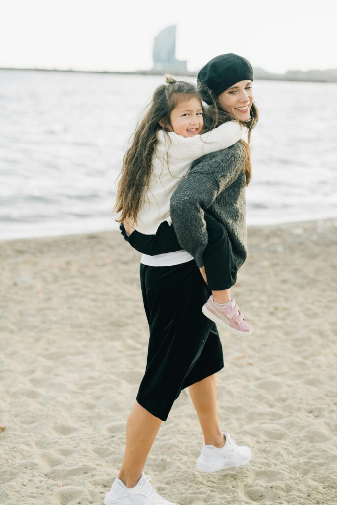 A mother gives her smiling daughter a piggyback ride on a deserted winter beach, with the W Hotel in the background.